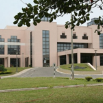 img_9505 - NubianBiz | African Business Directory, Jobs, Marketplace & Entrepreneur Insights Exterior view of a large beige modern building with tall glass windows and a curved driveway, surrounded by green lawn.